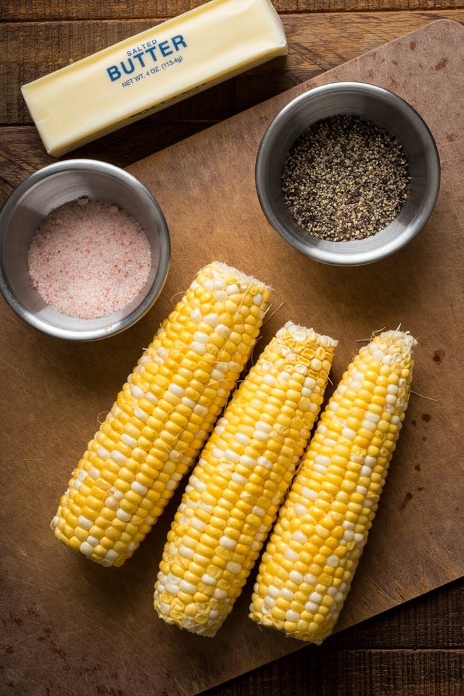 Fresh ears of yellow and white corn with salted butter, salt, and black pepper on a wooden cutting board.
