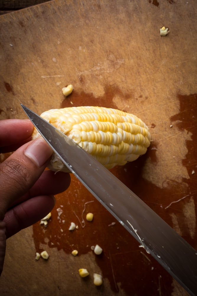Cutting fresh corn kernels from an ear of corn with a chef's knife on a wooden cutting board.